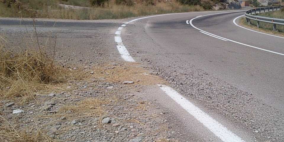 Detalle de una curva en carretera con el arcén y la línea exterior completamente cubiertos de gravilla y arena, ilustrando el peligro extremo de perder agarre en moto al trazar demasiado abierto.